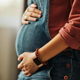Eine schwangere Frau h&auml;lt behutsam die H&auml;nde auf ihren Bauch und tr&auml;gt eine Jeans Latzhose